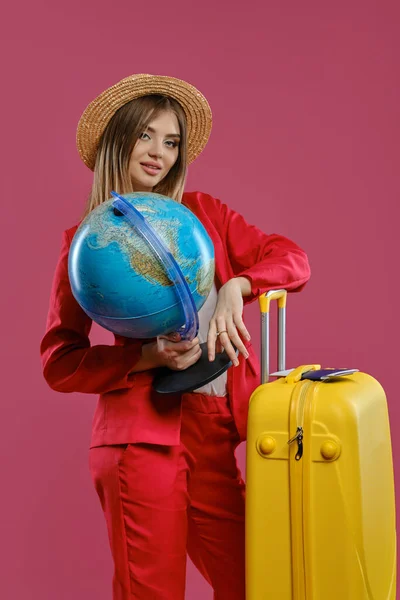 Модель в шляпе, белая блузка, красный брючный костюм. Улыбаясь, держа глобус. Опираясь на желтый чемодан, паспорт и билет на него, позируя на розовом фоне — стоковое фото