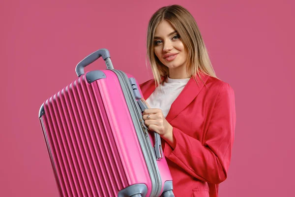 Blonďatý model v červeném kostýmu a bílé blůze. Usmívá se, drží kufr, pózuje na růžovém pozadí. Móda, cestování, reklama. Detailní záběr — Stock fotografie