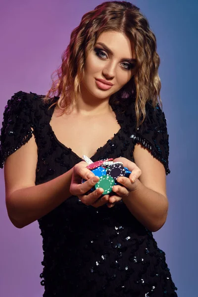 Blonde girl in black sequin dress is smiling, showing handful of multicolored chips, posing on colorful studio background. Poker, casino. Close-up.