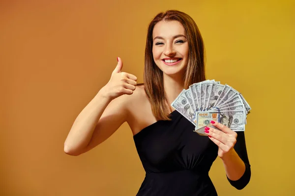 Blondynka w czarnej sukience uśmiecha się, pokazuje kciuk w górze, trzyma fankę studolarowych banknotów, pozuje na kolorowym tle. Poker, kasyno. Zbliżenie — Zdjęcie stockowe