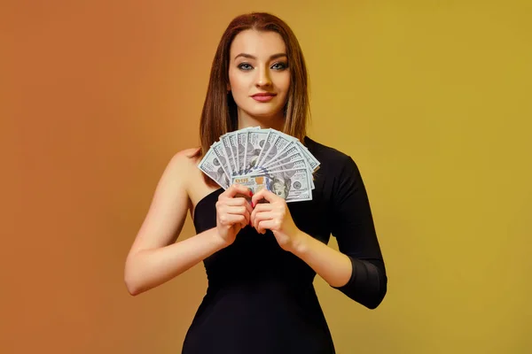 Blondynka w czarnej sukience uśmiecha się, pokazując fankę studolarowych banknotów, pozujących na kolorowym tle. Poker, kasyno. Zbliżenie. — Zdjęcie stockowe