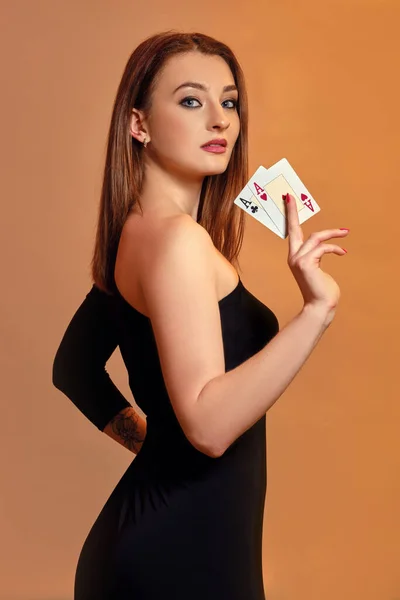 Ragazza bionda con trucco luminoso, in abito nero sta mostrando due assi, in posa lateralmente sullo sfondo colorato. Gioco d'azzardo, poker, casinò. Primo piano . — Foto Stock