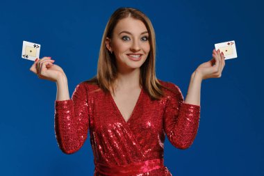 Makyajlı, kırmızı pullu elbiseli sarışın kadın gülümsüyor, iki as gösteriyor, mavi arka planda poz veriyor. Kumar, poker, kumarhane. Yakın plan..