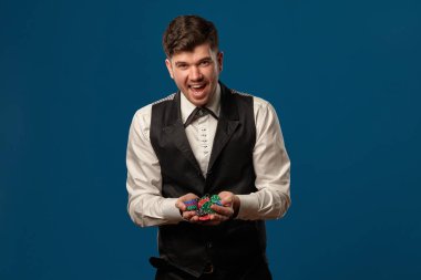 Pokerde çaylak, siyah yelek ve pantolon, beyaz gömlek. Renkli cips tutuyorum. Mavi arka planda poz vermek. Kumar, kumarhane. Yakın plan..