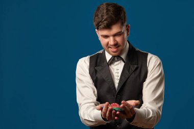 Pokerde çaylak, siyah yelekli ve beyaz gömlekli. Renkli cips tutuyorum. Mavi arka planda poz vermek. Kumar, kumarhane. Yakın plan..