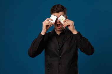 Pokerde acemi, siyah yelekli ve gömlekli. Mavi stüdyo geçmişine karşı poz verirken elinde iki iskambil kağıdı tutuyor. Kumar, kumarhane. Yakın plan..