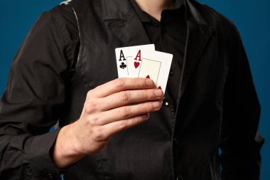 Pokerde çaylak, siyah yelekli ve gömlekli. Mavi stüdyo geçmişine karşı poz verirken elinde iki iskambil kağıdı tutuyor. Kumar, kumarhane. Yakın plan..