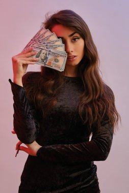 Siyah kadife elbiseli esmer kız iki kırmızı fiş ve biraz para gösteriyor, Coloful 'un arka planında poz veriyor. Kumar, poker, kumarhane. Yakın plan..