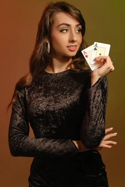 Bruneta dívka v černých sametových šatech a šperky se usmívá, ukazuje dvě esa, pózování na barevném pozadí studia. Hazard, poker, kasino. Detailní záběr — Stock fotografie