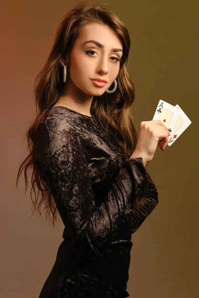 Barna hölgy fekete bársony ruhát és ékszereket mutat két ász, pózol oldalra színes stúdió háttér. Szerencsejáték, póker, kaszinó. Közelkép. — Stock Fotó