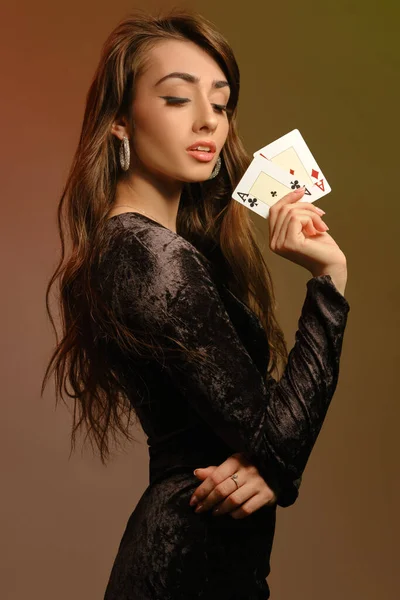 Bruneta žena v černých sametových šatech a špercích ukazuje dvě esa, pózuje bokem na barevném pozadí studia. Hazard, poker, kasino. Detailní záběr. — Stock fotografie