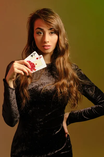 Bruneta žena v černých šatech ukazuje dva červené chipsy a esa, pózovat na barevném pozadí studia. Hazard, poker, kasino. Detailní záběr. — Stock fotografie
