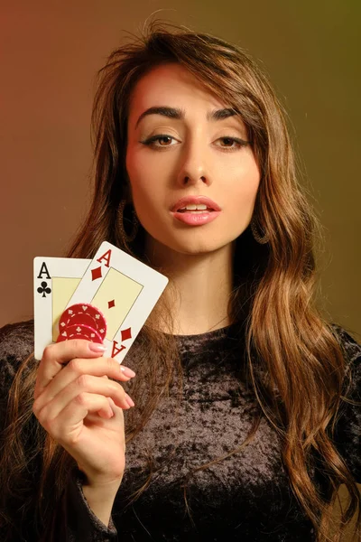 Brunette lady in black dress showing two red chips and aces, posing against colorful studio background. Gambling, poker, casino. Close-up. — Stock Photo, Image