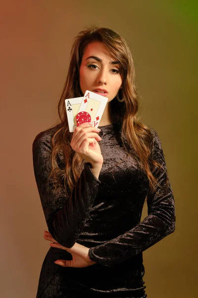 Bruna femmina in abito nero che mostra due chip rossi e assi, in posa contro lo sfondo colorato studio. Gioco d'azzardo, poker, casinò. Primo piano . — Foto Stock