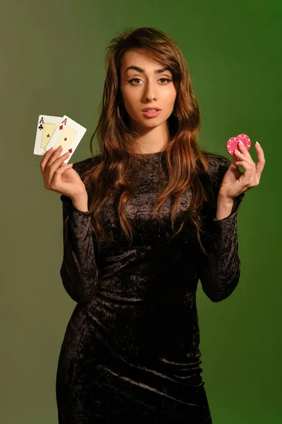 Bruneta dívka v černých šatech ukazuje dvě červené chipsy a esa, pózování na barevném pozadí studia. Hazard, poker, kasino. Detailní záběr. — Stock fotografie