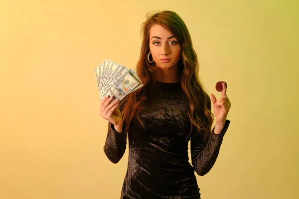 Brunetka dziewczyna w czarnej sukience pokazuje dwa czerwone żetony i trochę pieniędzy, pozowanie na żółtym tle studio. Hazard, poker, kasyno. Zbliżenie. — Zdjęcie stockowe