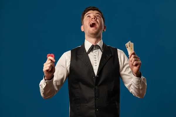 Noob im Poker, in schwarzer Weste und weißem Hemd. Mit zwei roten Chips und Assen. Posieren vor blauem Hintergrund. Glücksspiel, Casino. Nahaufnahme. — Stockfoto