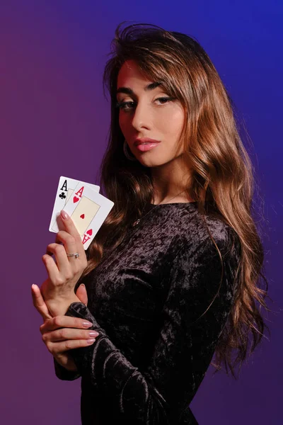 Bruneta dívka v černých sametových šatech ukazuje dvě hrací karty, pózování bokem na coloful pozadí. Hazardní hry, poker, kasino. Detailní záběr. — Stock fotografie