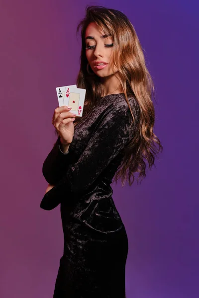 Brünettes Mädchen in schwarzem Samtkleid, das zwei Spielkarten zeigt und seitlich auf buntem Hintergrund posiert. Glücksspiel, Poker, Casino. Nahaufnahme. — Stockfoto