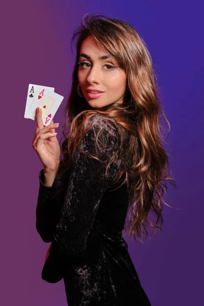Ragazza bruna in abito di velluto nero che mostra due carte da gioco, in posa lateralmente su sfondo colorato. Intrattenimento di gioco, poker, casino '. Primo piano . — Foto Stock