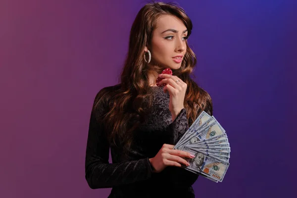 Bruneta dívka v černých sametových šatech ukazuje dva červené chipsy a nějaké peníze, pózování na pozadí Coloful. Hazard, poker, kasino. Detailní záběr. — Stock fotografie