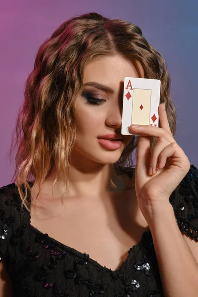 Hnědovlasá dívka v černých lesklých šatech si zakryla oko diamantovým esem, pózuje na barevném pozadí. Hazard, poker, kasino. Detailní záběr. — Stock fotografie