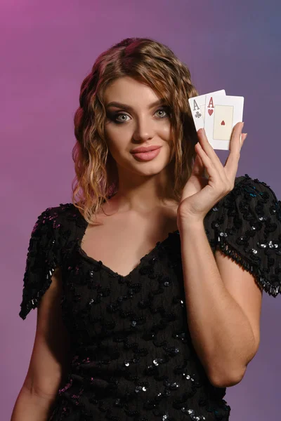 Braunhaarige Mädchen in schwarzem, glänzendem Kleid lächeln, zeigen zwei Asse und posieren auf buntem Hintergrund. Glücksspiel, Poker, Casino. Nahaufnahme. — Stockfoto