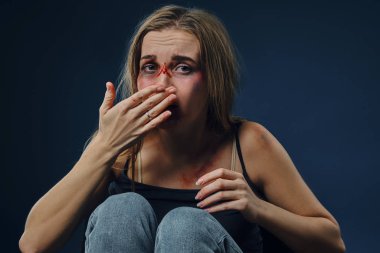 Siyah tişörtlü ve kot pantolonlu korkmuş kız. Yüzü morluklarla kaplıydı, burnuna dokunuyordu, mavi arka planda oturuyordu. Yakın plan.