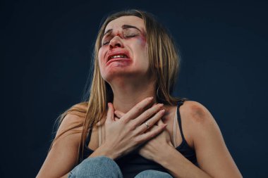 Siyah tişörtlü ve kot pantolonlu korkmuş bir kız. Yüzü morluklarla kaplıydı, ellerini göğsüne koydu ve ağladı, mavi arka planda oturuyordu. Yakın plan.