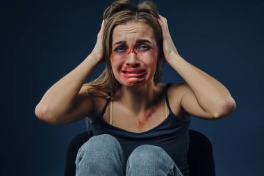 Korkmuş bir kadın, yüzü morluklarla kaplı, siyah tişört ve kot pantolon giymiş, başını tutup ağlayan, mavi arka planda oturan. Yakın plan.