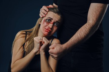 Zalim adam elini kadının kafasına koydu. Yüzünde morluklar olan korkmuş bir kurban yakınlarda oturuyor, korkmuş. Mavi arka plan. Aile içi şiddet. Yakın plan.