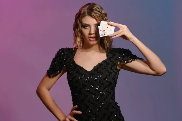 Hnědovlasá dáma v černých lesklých šatech má zakryté oko se dvěma hracími kartami, pózující na barevném pozadí. Hazard, poker, kasino. Detailní záběr. — Stock fotografie