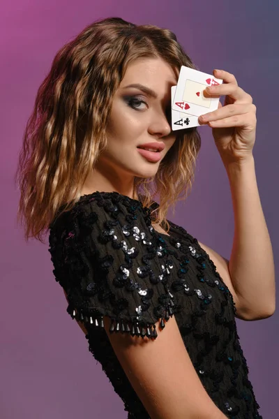 Καστανά μαλλιά κορίτσι σε μαύρο γυαλιστερό φόρεμα δείχνει δύο άσους, ποζάροντας πλάγια σε πολύχρωμο φόντο. Ψυχαγωγία, πόκερ, καζίνο. Κοντινό πλάνο. — Φωτογραφία Αρχείου
