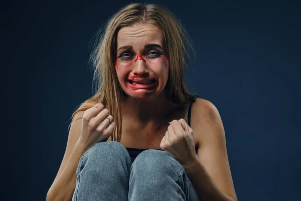 Asustada doncella en camiseta negra y jeans. Cara sangrante cubierta de moretones, apretó los puños y lloró, sentada sobre fondo azul. Primer plano —  Fotos de Stock