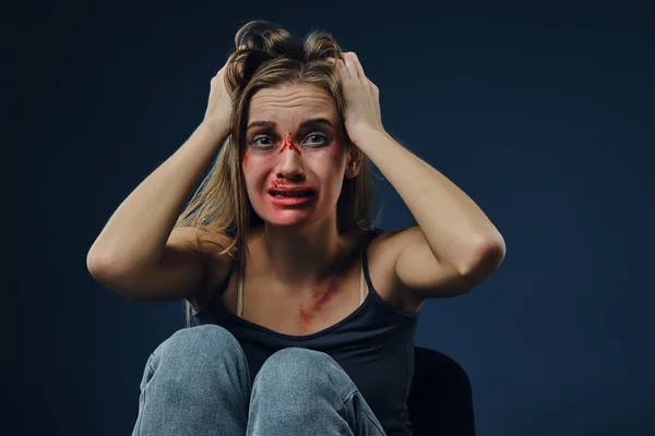 Scared female, bleeding face covered with bruises, in black t-shirt and jeans, grabbed own head and crying, sitting on blue background. Close-up