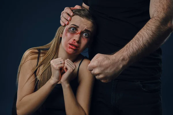Kegyetlen férj rátette a kezét a feleségére. Félős áldozat, zúzódásokkal az arcán, rémülten ül a közelben. Kék háttér. Családi erőszak. Közelkép — Stock Fotó