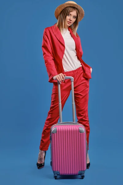 Blondynka w słomkowym kapeluszu, biała bluzka, czerwony garnitur, wysokie czarne obcasy. Opiera się na różowej walizce, pozując na niebieskim tle. — Zdjęcie stockowe