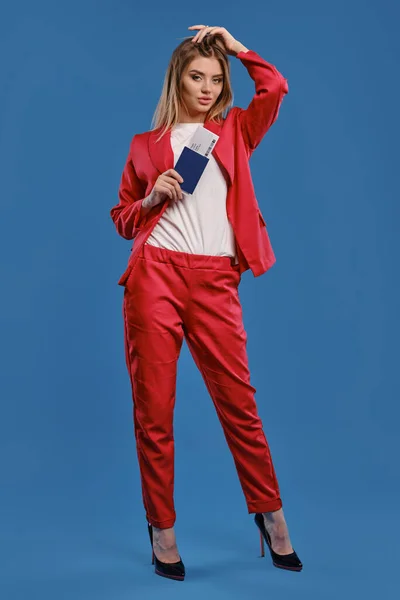 Блондинка в белой блузке, красный брючный костюм, высокие черные каблуки. Она трогает волосы, держит паспорт и билет, позирует на синем фоне. Полная длина — стоковое фото