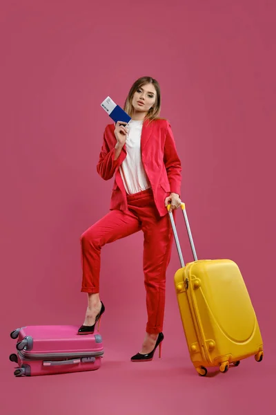 Mulher loira de fato vermelho, blusa branca, salto preto. Ela colocou a perna na mala, amarela por perto, segurando passaporte e bilhete, fundo rosa — Fotografia de Stock