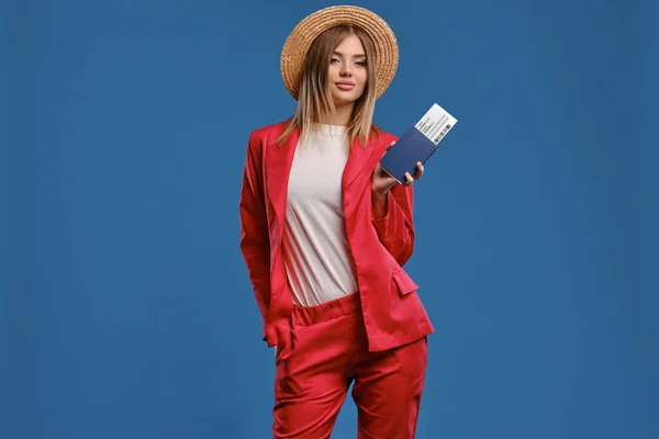 Блондинка в соломенной шляпе, белой блузке и красном брючном костюме. Она улыбается, держа в руках паспорт и билет, позируя на фоне синей студии. Крупный план — стоковое фото