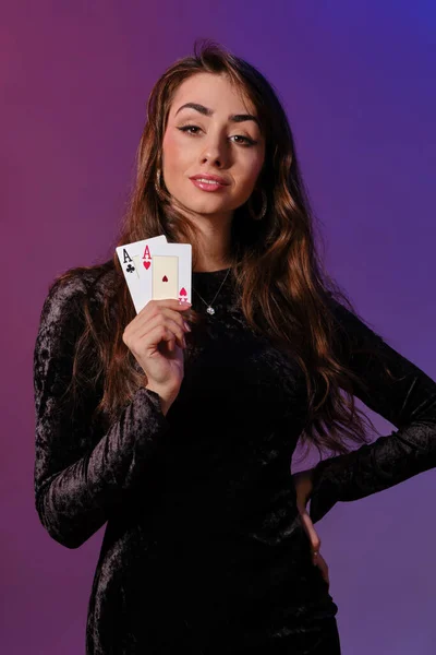 Brunett kvinna i svart sammet klänning visar två spelkort, poserar mot coloful bakgrund. Spelunderhållning, poker, kasino. Närbild. — Stockfoto