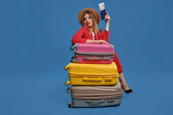 Блондинка в соломенной шляпе, белая блузка, красный брючный костюм. Сидя, опираясь на красочные чемоданы, держа в руках паспорт и билет, позируя на синем фоне — стоковое фото