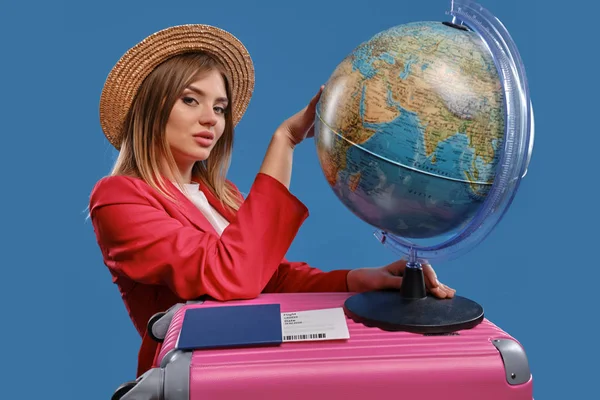 Blonde female in straw hat, white blouse, red jacket. Holding globe standing on pink suitcase, passport and ticket nearby, posing on blue background — Stock Photo, Image