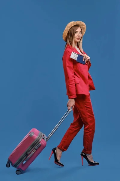 Блондинка в шляпе, белая блузка, красный брючный костюм, высокие каблуки. С розовым чемоданом, с паспортом и билетом, позируя боком на синем фоне — стоковое фото