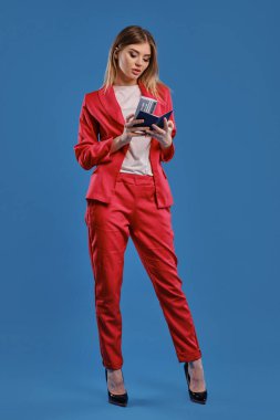 Blonde lady in white blouse, red pantsuit, high black heels. She is looking at her passport and ticket while posing on blue background. Full length