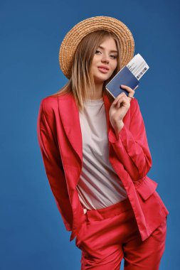 Blonde girl in straw hat, white blouse and red pantsuit. She smiling, showing passport and ticket while posing on blue studio background. Close-up