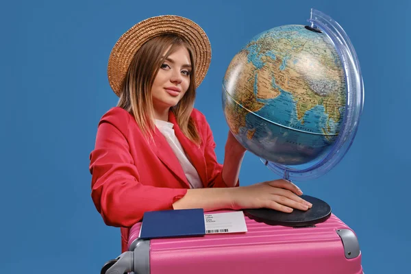 Blondes Mädchen mit Strohhut, weißer Bluse, roter Jacke. Globus auf rosa Koffer, Pass und Ticket in der Nähe, posierend auf blauem Hintergrund — Stockfoto