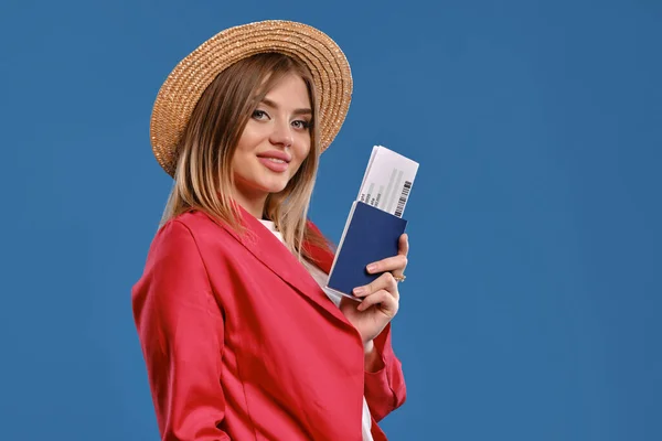 Блондинка в соломенной шляпе, белая блузка, красный брючный костюм. У нее есть паспорт и билет, позирует боком на синем фоне. Путешествую. Крупный план — стоковое фото