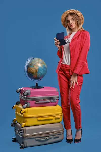 Mulher de chapéu, blusa branca, fato vermelho, saltos pretos. Ela está segurando passaporte e bilhete, posando em fundo azul. O globo está em cima das malas. — Fotografia de Stock