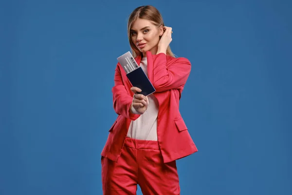 Blonde lady in white blouse and red pantsuit. She is tucking her hair, holding passport and ticket while posing on blue studio background. Close-up — 스톡 사진